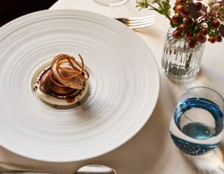 A coffee and cacao dessert with a chocolate quenelle and crisp garnish, served on a white dish with concentric grooves.