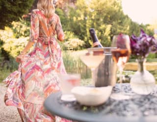 A woman in a flowing dress enjoys the garden. A posy of Sweet Peas stands in a vase, beside a silver bucket of champagne