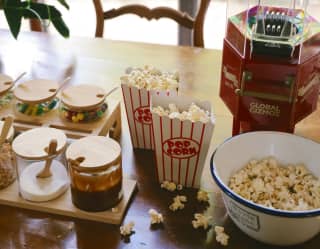 A old-style popcorn maker with two full cinema buckets and a bowl sit on a table alongside jars of toppings for movie night.