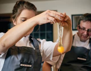 Girl in chef whites cracking an egg as a teaching chef looks on