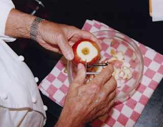 A chef uses a peeler to skin an apple, collecting peel in a glass bowl atop a red and white checked cloth, seen from above.