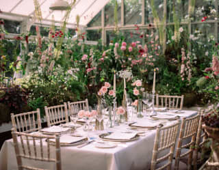 A laid table with pink roses and candles waits for guests in the magical Hartley botanic glasshouse, filled with hollyhocks.