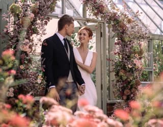 A bride and groom stand below an arch of pink flowers that surrounds the doorway of the elegant Victorian glasshouse