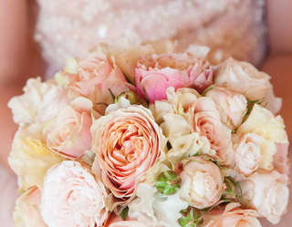 A close up image of a bride holding a round bouquet of roses in pastel shades of pink, peach and cream