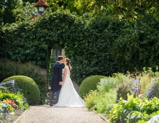 Bride and groom sharing a kiss under an arch in a leafy hedge along a garden path