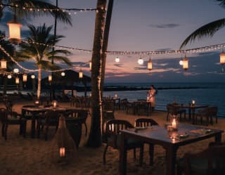 Lanterns and light strings criss-cross the darkening sky above dining tables on the sand, as a waitress lights candles.