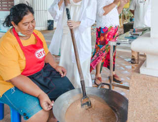 A local woman in a white dress shows a guest how to make Kalamay, mixing coconut milk, brown sugar and ground rice in a pan.