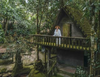 A woman stands on the mossy balcony of a little stone house in the Secret Buddha Garden, set in lush jungle.