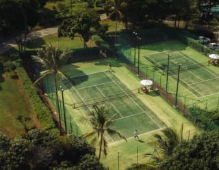 An aerial shot of a man playing tennis on one of two dark green courts with lime grass surrounds and white parasol shades.