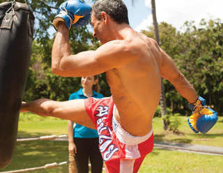 Gentleman in red boxing shorts and blue boxing gloves high-kicking a punchbag