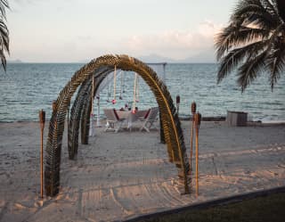 A table facing the ocean waits for a honeymooning couple, seen through a row of frond-shaped arches, set into the beach sand.
