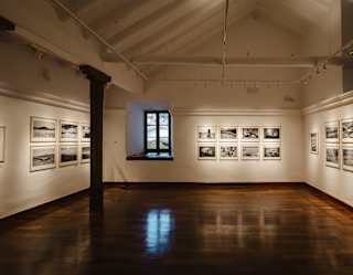 An art gallery with wooden floors and white walls displays framed black-and-white photographs. A window is centred on the back wall, letting in natural light. The room is brightly lit with spotlights above.