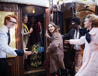 A steward in white coat with gold epaulettes greets travellers as they rush to join their friends and board the train