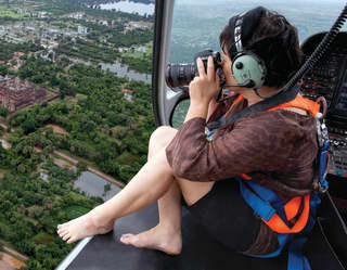 Photographer perching on the edge of a helicopter soaring over Angkor Wat
