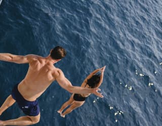 Action shot from above of a man leaping from a height following a woman forming a dive - both in mid-air with the sea below.