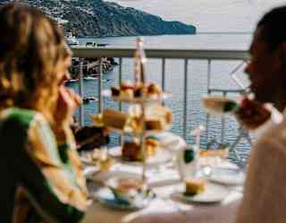 A couple in soft-focus foreground enjoy tiers of treats for Afternoon Tea at a terrace table overlooking the coast and ocean.