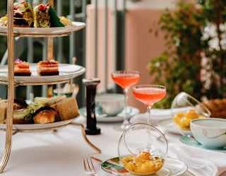 Eye-level view of an Afternoon Tea, with a cake stand of sweet and savoury treats, two pink drinks and glass bowls of fruit.