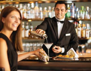 A female guest at the bar in a black dress with a gold purse smiles as a bartender pours her a cocktail from a silver shaker.