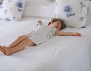 A smiling young child in short dungarees lies on their back with arms spread on a fresh white bed with floral cushions.