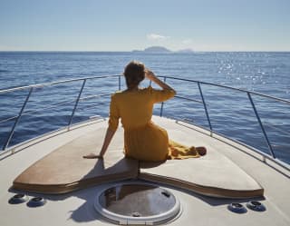 Lady in a yellow dress sitting on the bow of a sailing boat heading towards islands