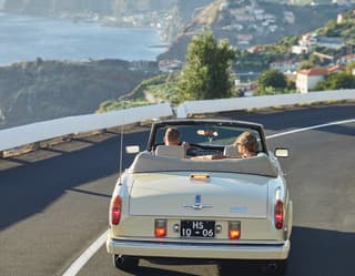 Lady and a driver in an open-top classic car driving along a coastal road
