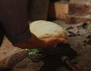 A man lifts a large boule of raw dough towards an open brick oven, seen from behind in low light, close-up.