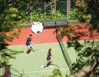 Seen through trees, a mixed pair play tennis on a sunny green and orange court, with a refreshments table on the side line.
