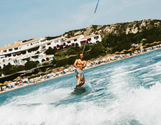 A man mono-skis in the waters in front of Romazzino hotel, seen from the back of a boat with the tow rope passing overhead.