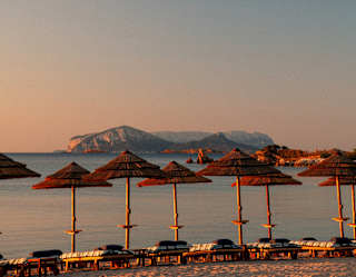 Sun glows on thatched parasols and flat sun-loungers lining the waterfront as island contours rise into a soft sunset sky.