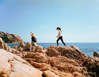 Two women - one blond, one dark - walk across the brown bumpy granite rock formations along the coast with sea behind.