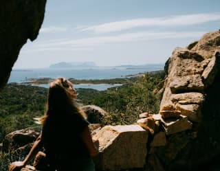 Sitting on a stone wall in the hills, a woman gazes over forest towards Pevero Beach and islands beyond, viewed from behind.