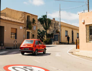 In San Pantaleo, a 1970s red Fiat 126 crosses a road painted with a circular red and white 30-kilometre speed limit sign.