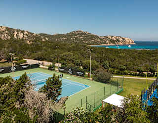 A rocky headland and flashes of bright azure ocean are seen behind a coastal forest, in a view from above the tennis court.