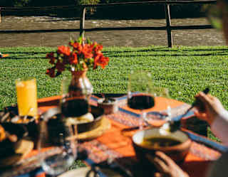 In soft-focus foreground a guest enjoys lunch and red wine at an El Jardin riverside table with an orange tablecloth.