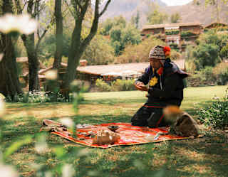A shaman in a traditional chullo hat kneels on a red-patterned blanket laid on the lawn and covered with shells and stones.