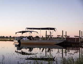 A flat-bottomed boat with a black canopy is moored on calm water at sunset. The sky is clear, and vegetation and trees line the horizon, reflected in the water’s surface.