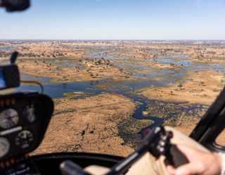 Aerial view from inside a helicopter cockpit flying over a vast, patchy wetland landscape with scattered trees, water channels, and dry areas under a clear blue sky.