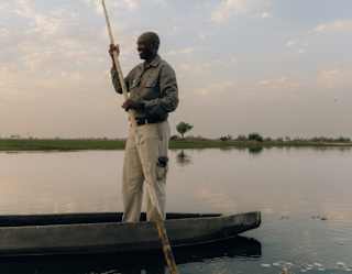 A tall mokoro safari guide smiles as he stands in a canoe holding a long pole which dips into the waterway, creating ripples.