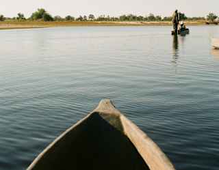A canoe glides on calm water under a clear sky, with another person standing on a boat in the distance, near a tree-lined shore.