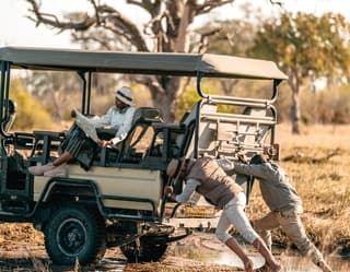 Two guests pushing a safari truck while a lady sits on top reading a map