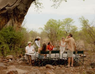 Three men and three women enjoy lunch on fold-out tables and chairs on the stone-studded earth beneath a giant baobab tree.