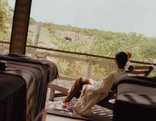 A man in a white robe and slippers reclines in a lounger on the deck of his accommodation, gazing at a passing elephant.