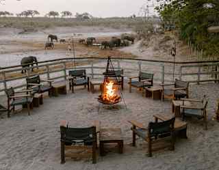 A hanging fire bowl with flaming logs provides the central focus in a sandy boma enclosure, circled by chairs and tables.