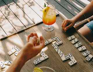 A couple enjoys cocktails and a game of dominos at a small wooden table on the deck, seen from above in close-up.