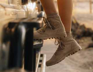 A woman with bare legs, wearing beige safari shoes, mounts a game viewing jeep in a close-up taken from the knees down.