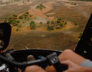 View of the Savannah in patches of parched brown and russet red, studded with trees, seen from a helicopter cockpit.