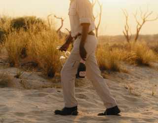 A man walks across sunset dunes with a Champagne bottle in one hand and two glasses in the other, seen from shoulders down.