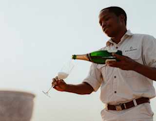 A male member of staff carefully pours Champagne from a bottle into a flute for a sundowner experience, seen at a low-angle.