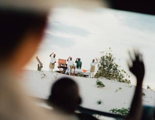 Four staff members, next to a table at the top of a white sand hill, wave to guests, seen through heads in the tourist jeep.