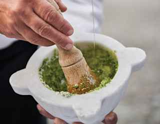 A chef drizzles olive oil from a small glass jug into a pestle and mortar, bright green with freshly pounded pesto ingredients